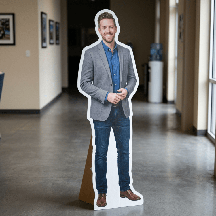 Life-size cardboard cutout of a man wearing a gray blazer, blue shirt, blue jeans, and brown shoes standing in an office hallway